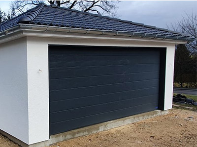 Auf dem Foto ist eine moderne Garage mit individuell geplantem Dach und Architektur zu sehen, die von der M.S. Garagenbau GmbH im Kundenauftrag sorgfältig geplant wurde und mit ihren blauen Dachziegeln perfekt an die Architektur des Hauses angepasst wurde.