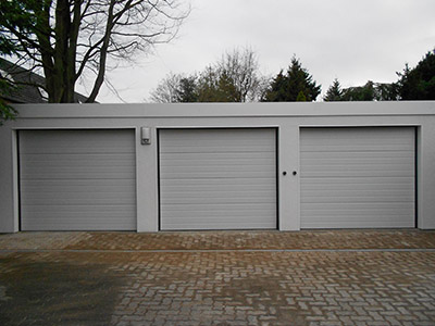 Maßgeschneiderte Reihengaragen: Effizienter Garagenbau für mehrere Fahrzeuge. Planen Sie Ihre individuellen Reihengaragen mit M.S. Garagenbau GmbH. Auf dem Foto sieht man eine einfache und dennoch effiziente Reihenanlage mit drei Garagen, welche Ton in Ton gestaltet worden sind: Wände, Dach und Rolltor sind in zeitlosem grau gehalten.