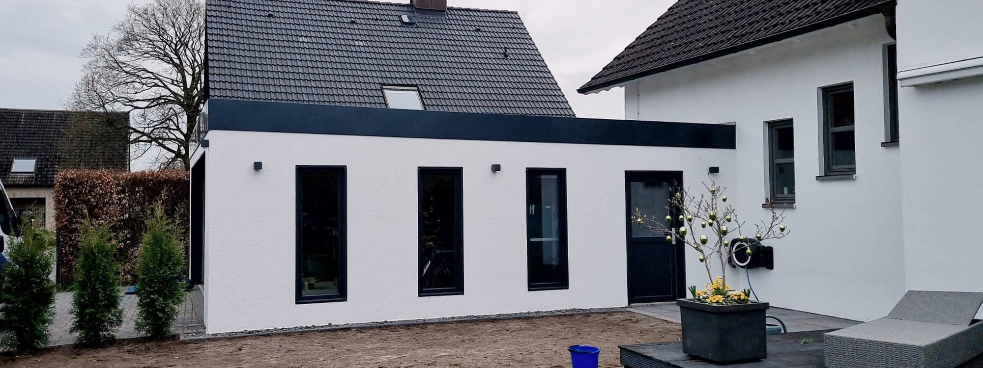Auf dem Bild ist eine moderne Großraumgarage zu sehen, die individuell geplant wurde, damit sie direkt an das angrenzende Haus anschließen kann. Die Garage ist weiß verputzt und hat blaue Dachziegel.