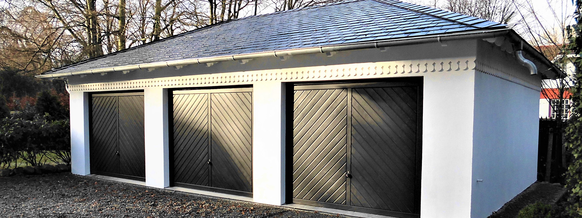 Das Bild zeigt eine moderne Reihengarage mit drei Toren für drei Fahrzeuge. Die Garage wurde weiß verputzt und mit einem dunklen, sehr elegantem Dach gebaut.